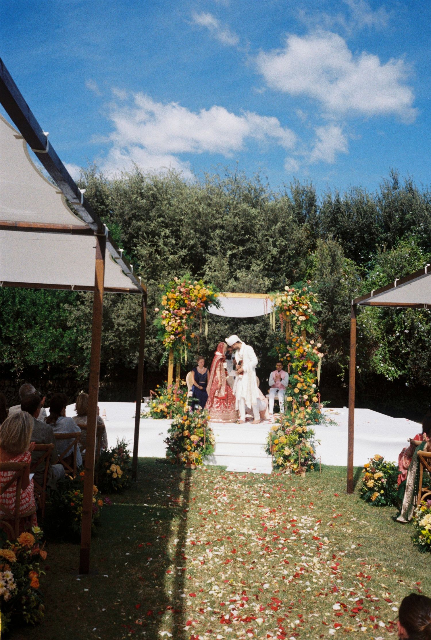 Outdoor wedding ceremony setup in an orchard with wooden posts, floral arrangements and scattered rose petals on the grass.