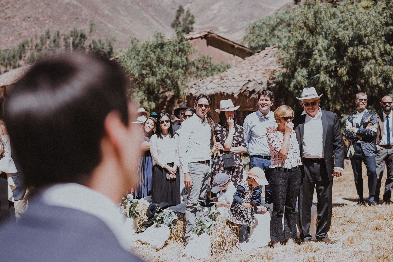 Wedding guests gather outdoors with mountains and rustic buildings in background.