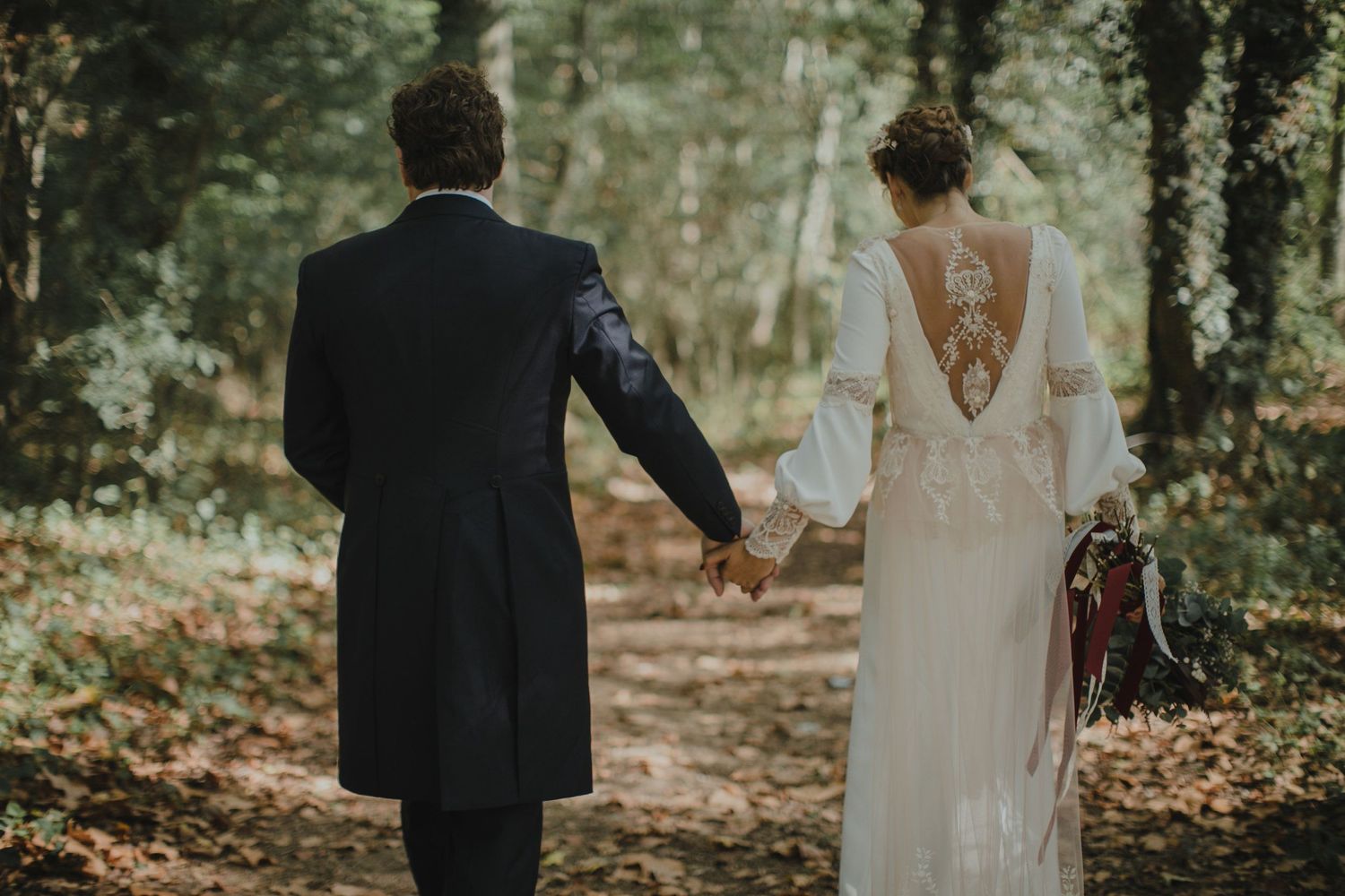 Elegant wedding dress with open back design as couple walks through wooded path.