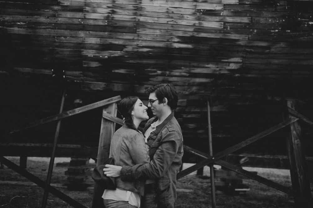 A couple embraces in a moody black and white setting under wooden beams.