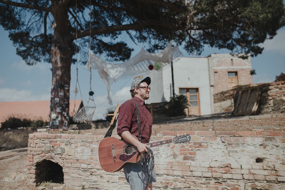 Musician with acoustic guitar performing outdoors against a rustic stone wall backdrop.