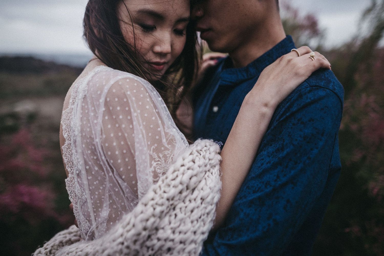 A romantic couple embraces in an intimate moment wearing white lace and blue against a moody outdoor backdrop.
