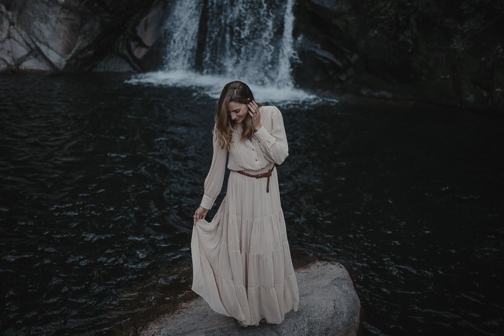 A person in a flowing beige dress stands gracefully on a rock in front of a cascading waterfall.