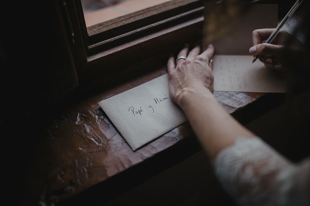 Close-up of hands writing on paper near window, wearing delicate lace bridal sleeve.