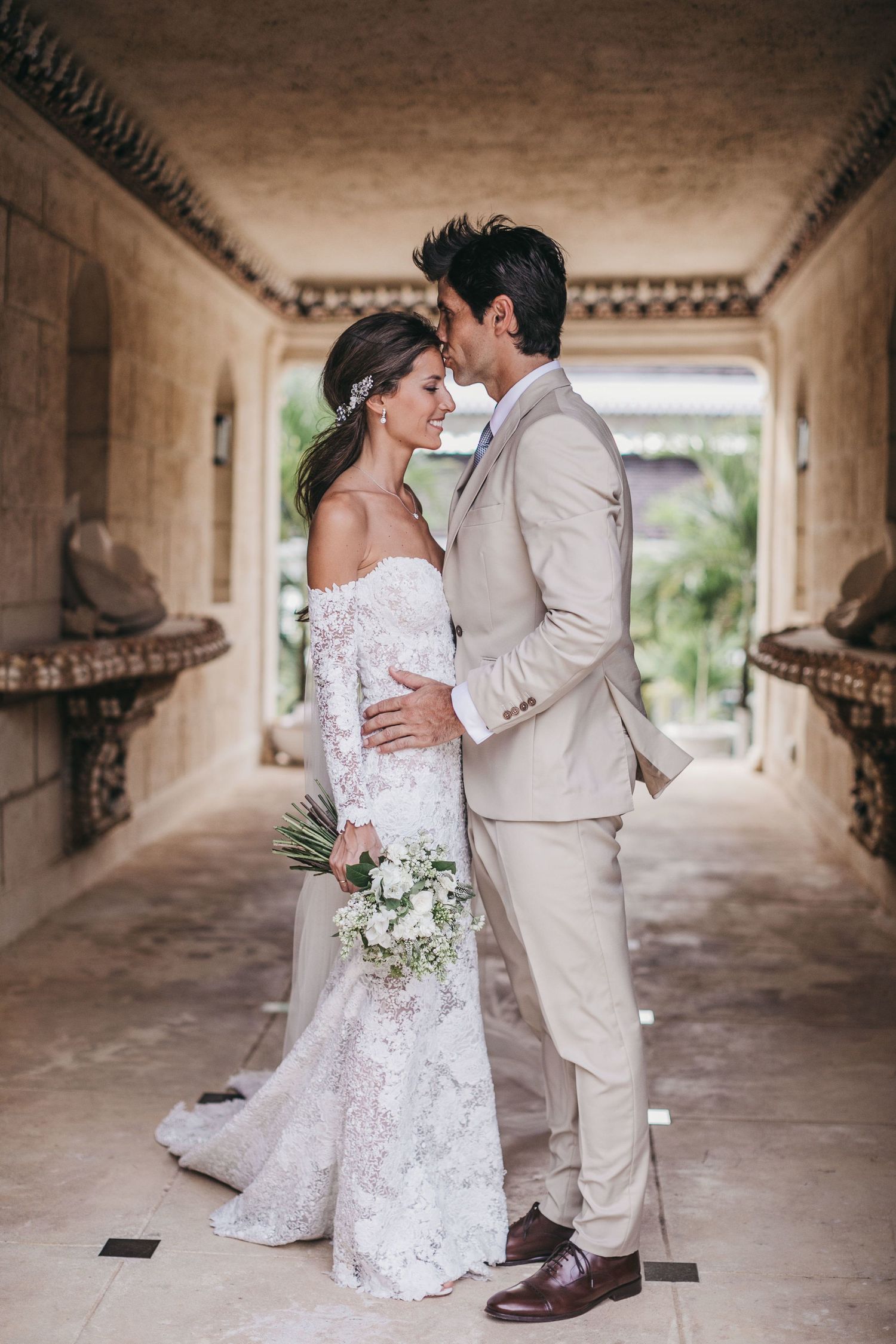 A romantic wedding couple embraces in an elegant corridor with off-white walls and decorative trim.