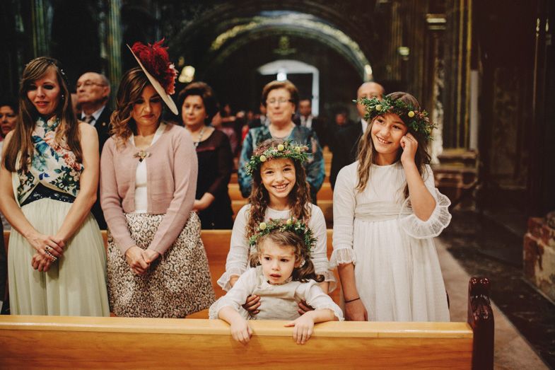 Young flower girls and attendants in white dresses seated in church pews during wedding ceremony.