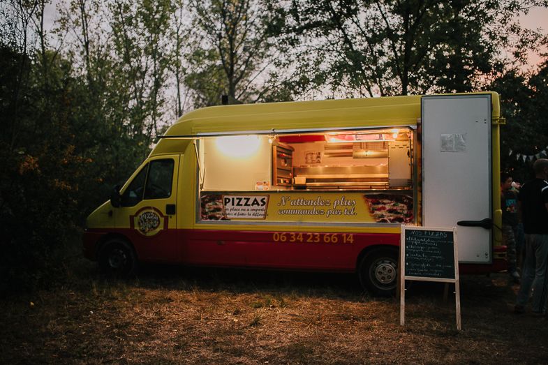 An illuminated yellow food truck serving pizza at dusk in a wooded area.