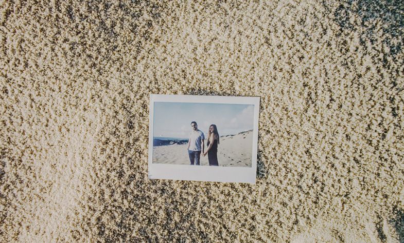A polaroid photo lying on sandy textured surface shows two figures at the beach.