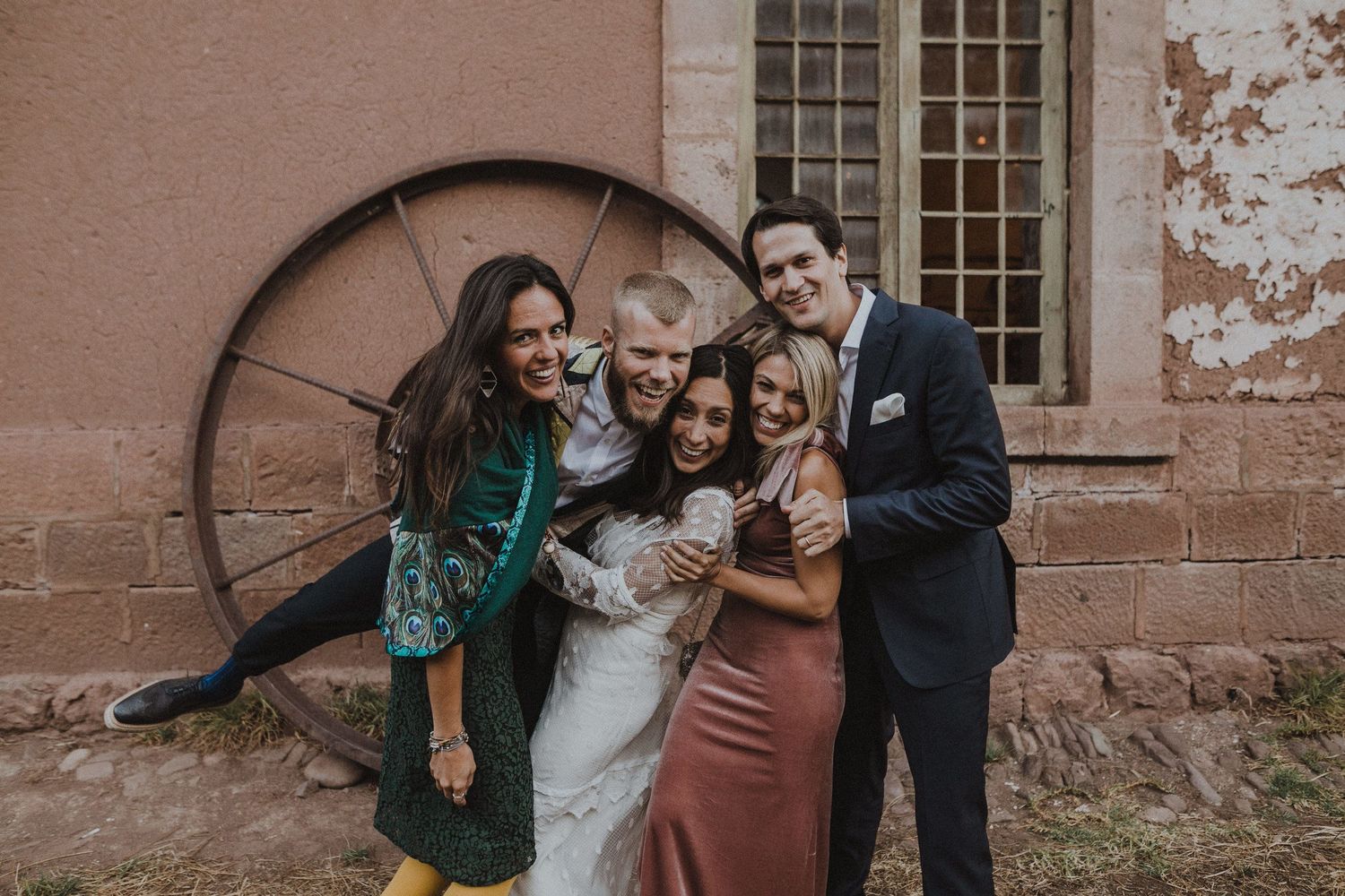 Wedding party poses together against rustic stone wall with vintage wagon wheel in background.