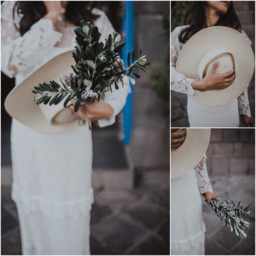 Elegant bride in lace dress holds greenery and a cream-colored hat against stone wall backdrop.