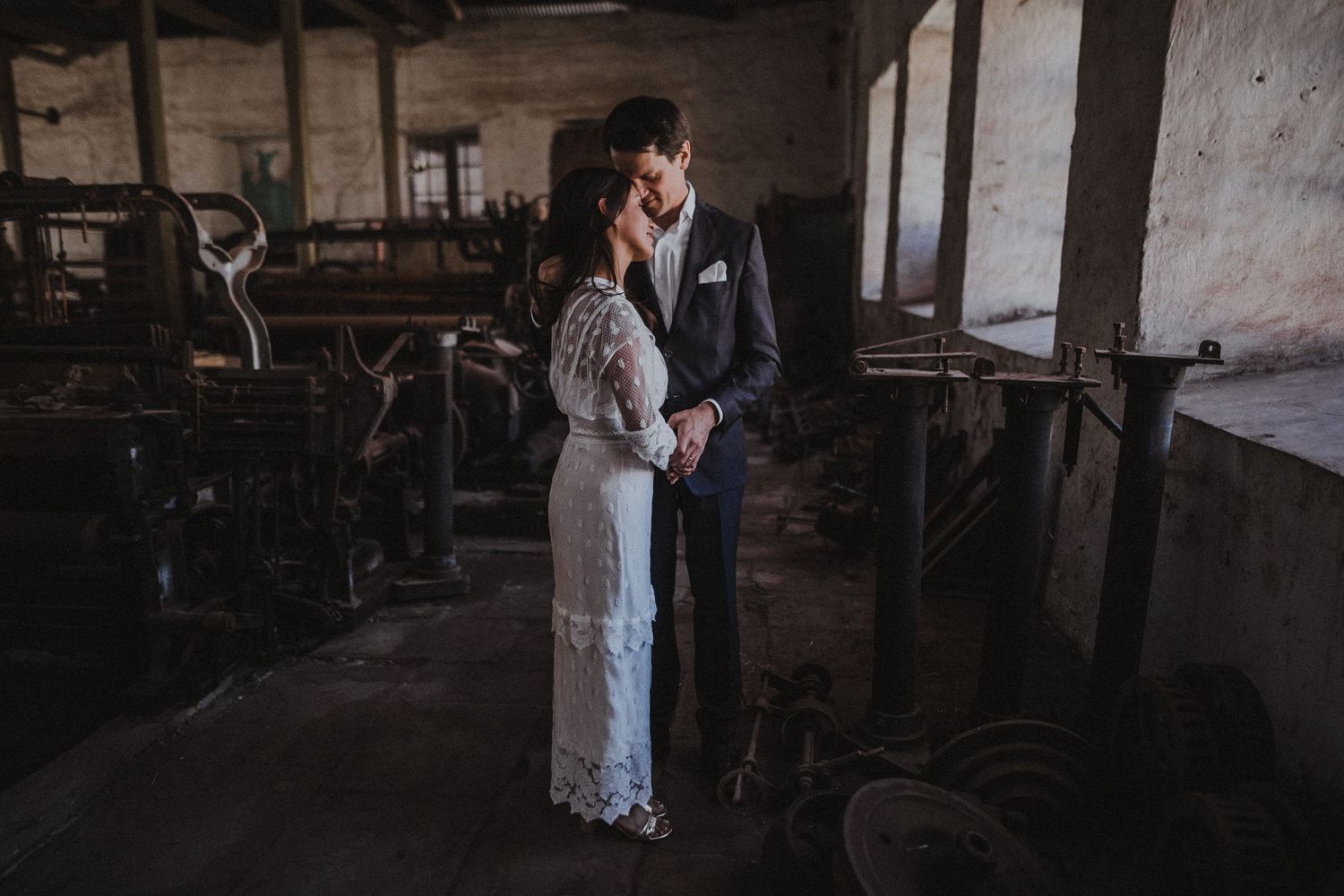 Couple in wedding attire embracing in a dimly lit industrial warehouse space.