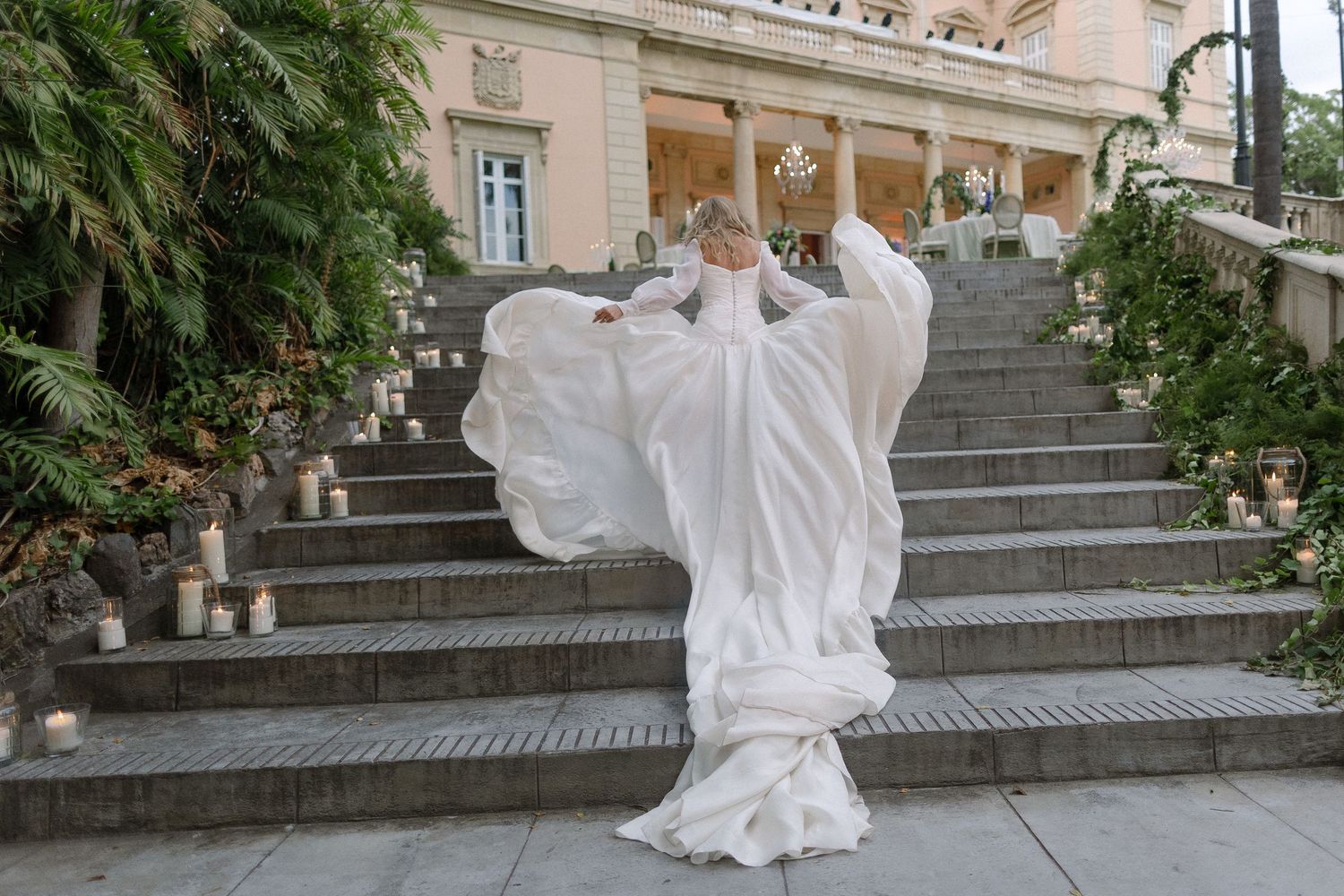 Bride in flowing white dress poses dramatically on grand stone staircase surrounded by greenery.