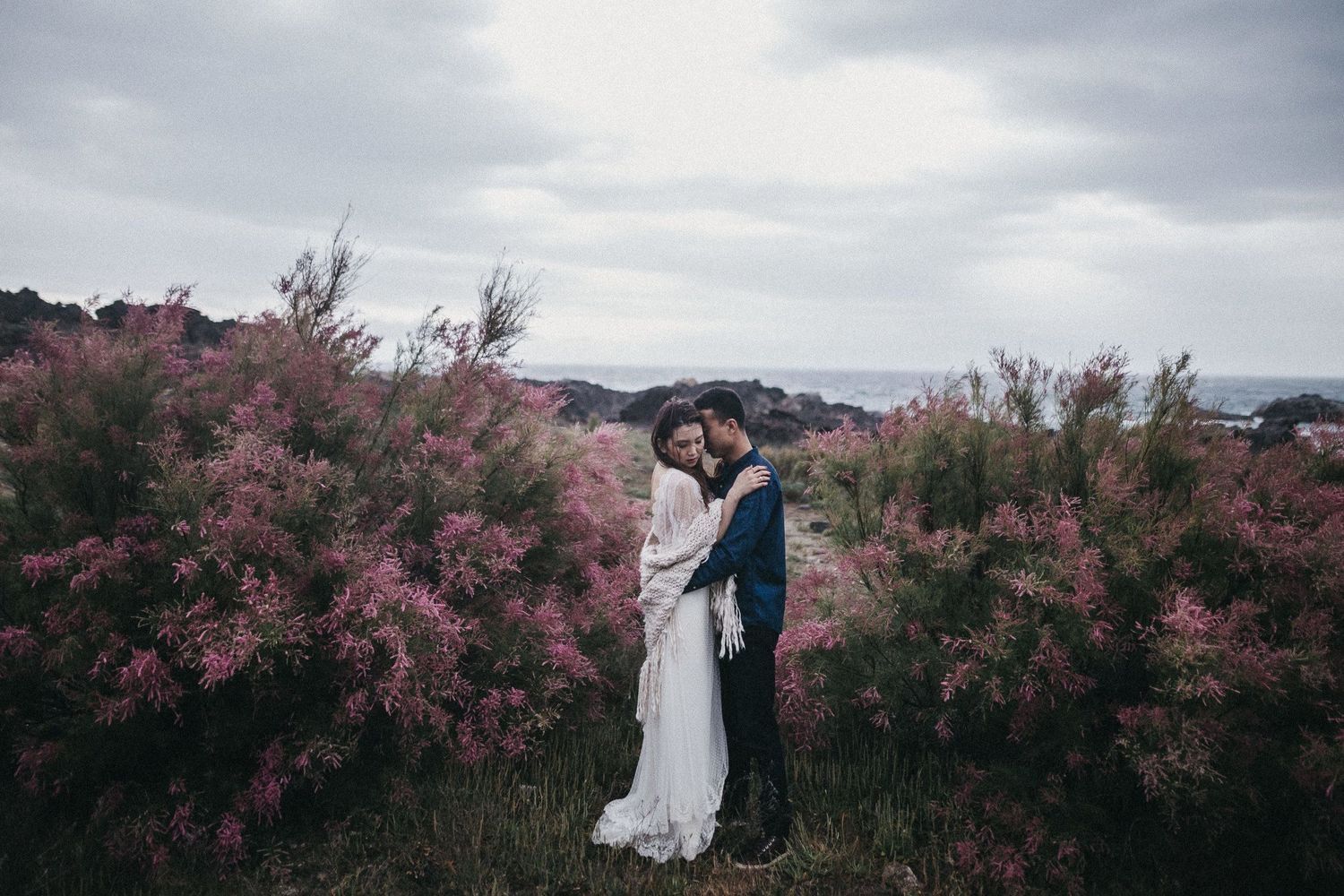 Couple in wedding attire standing among blooming pink flowers with cloudy sky backdrop.
