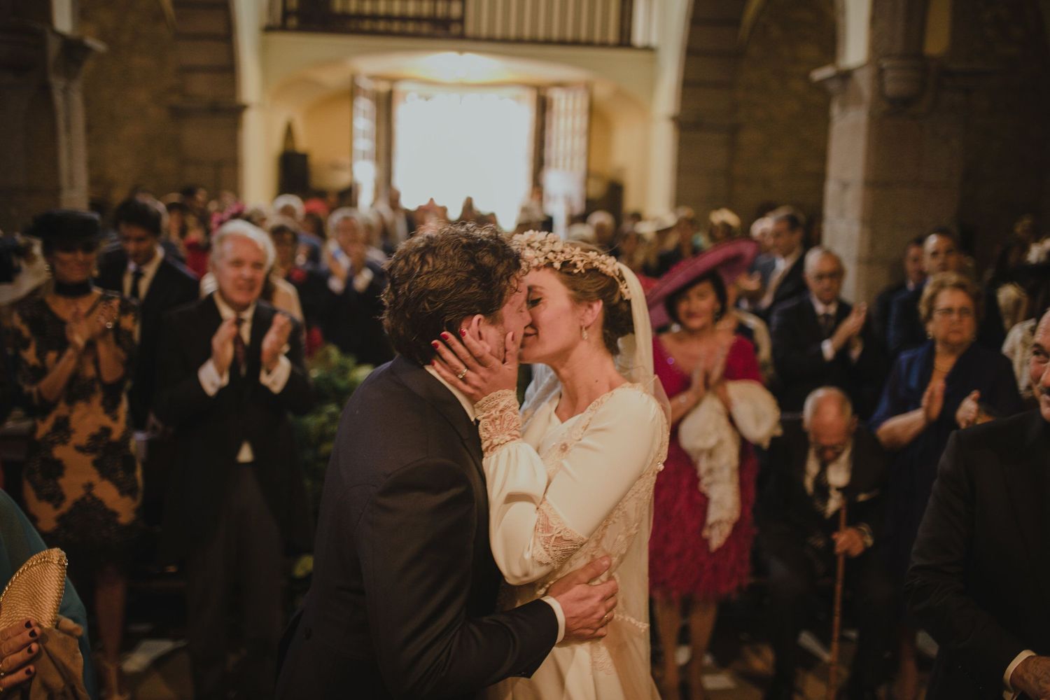 Wedding couple shares first kiss surrounded by applauding guests in historic stone church.