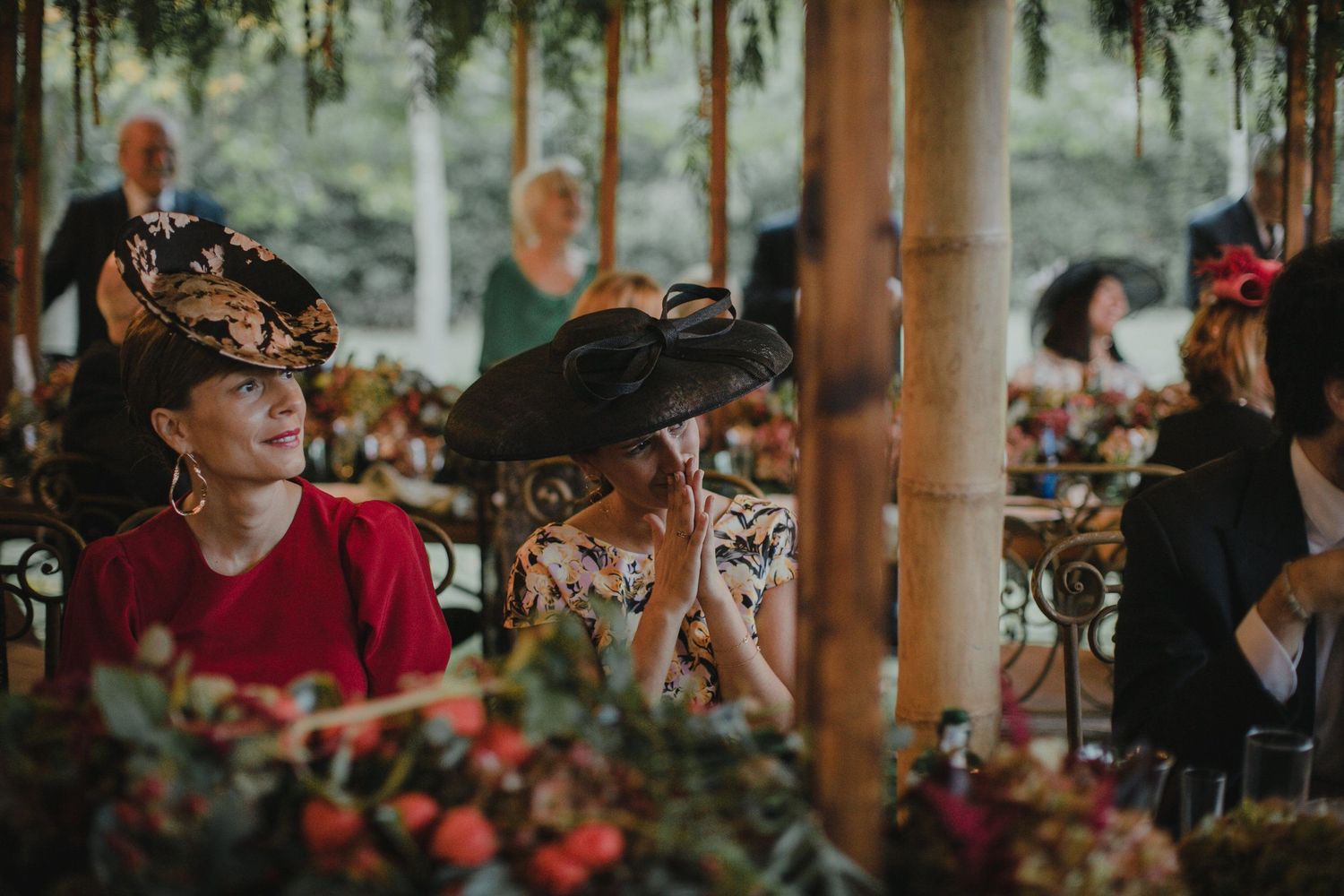 Elegant guests wearing decorative hats at an outdoor wedding reception under a covered venue.
