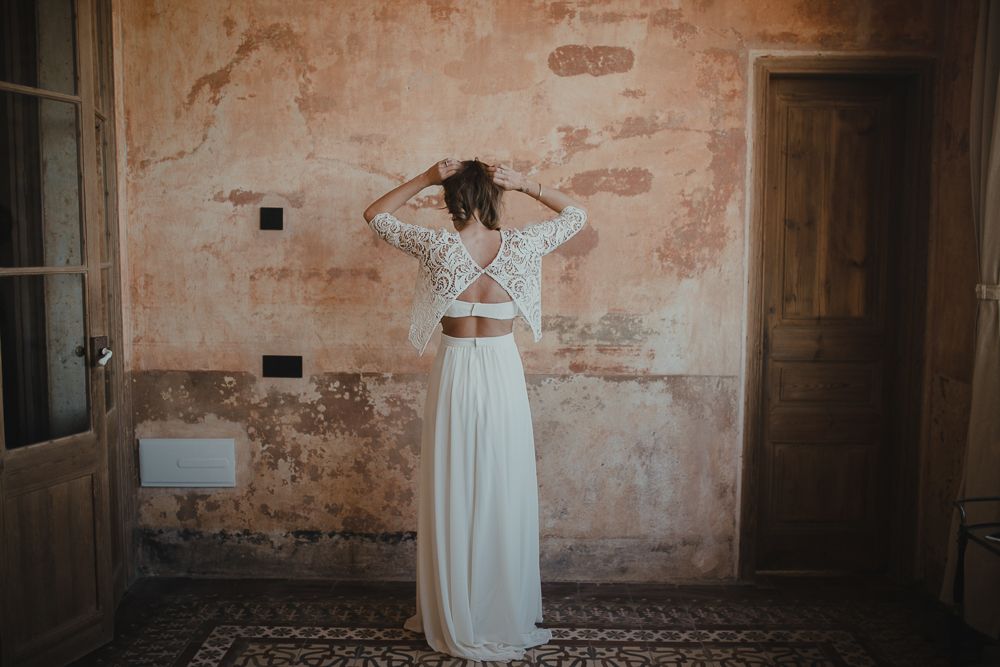 Back view of a two-piece wedding dress with lace top and flowing skirt against a rustic wall.