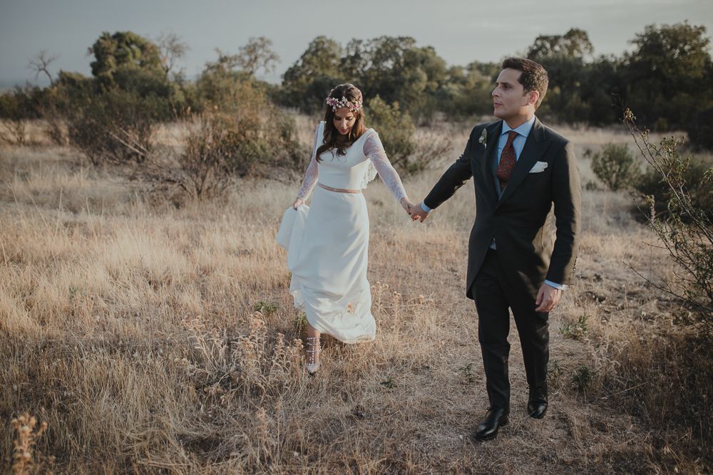 A couple walk hand in hand through a desert landscape during a wedding photoshoot.