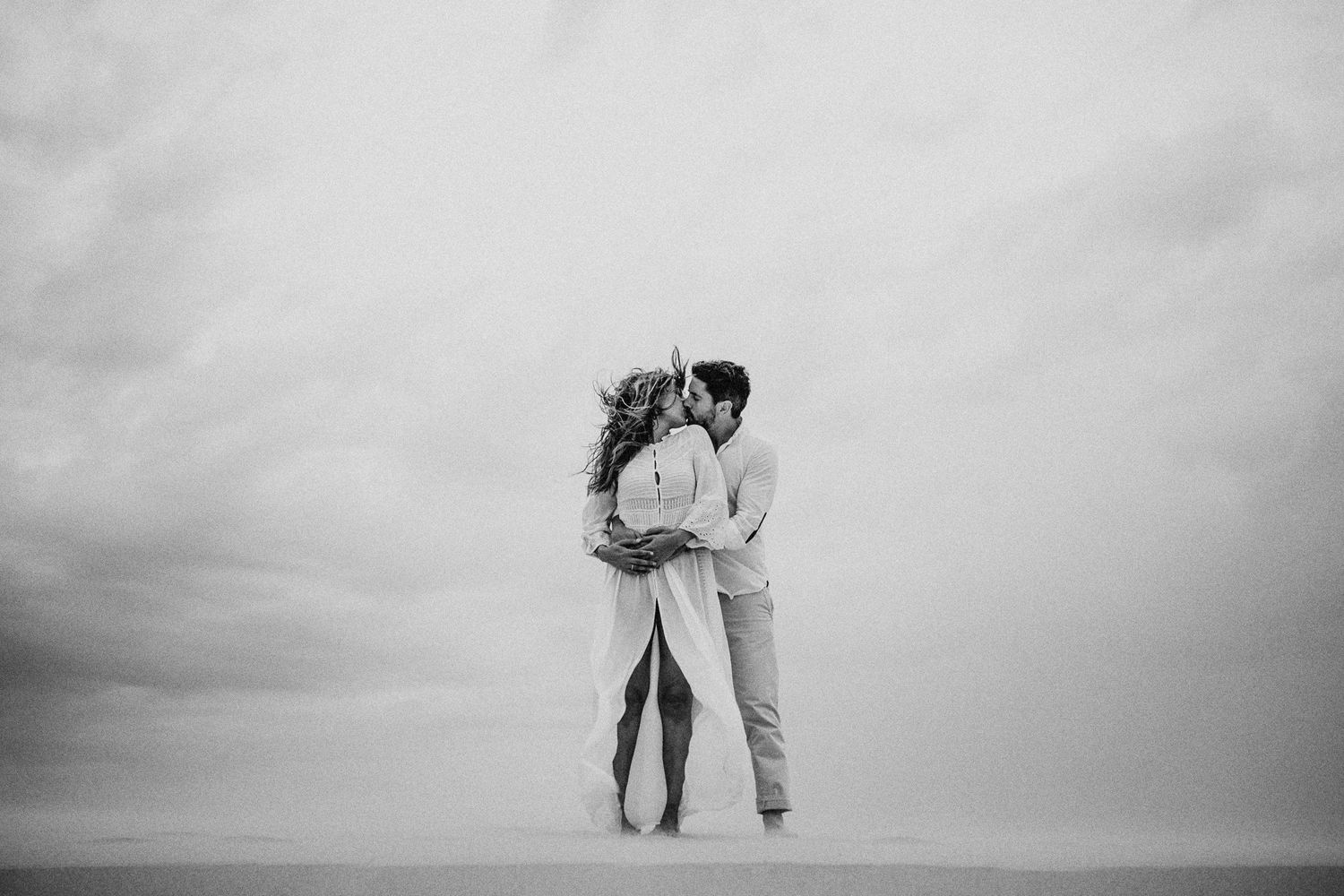 Black and white beach portrait of couple embracing against dramatic cloudy backdrop.