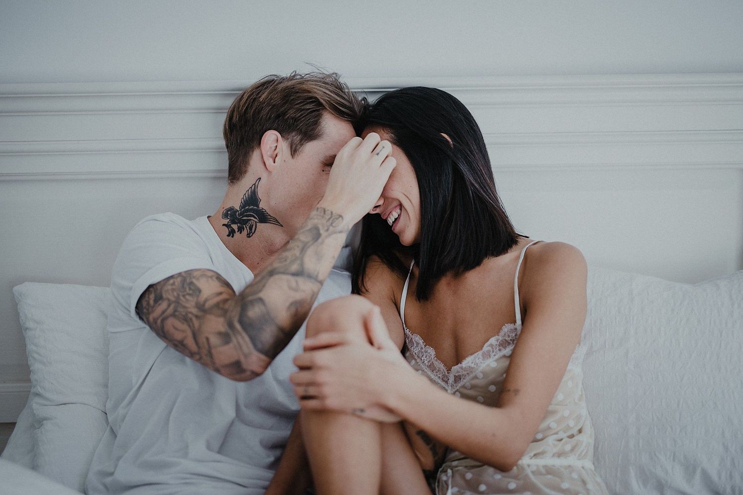 Couple laughing and embracing intimately on white bedding.