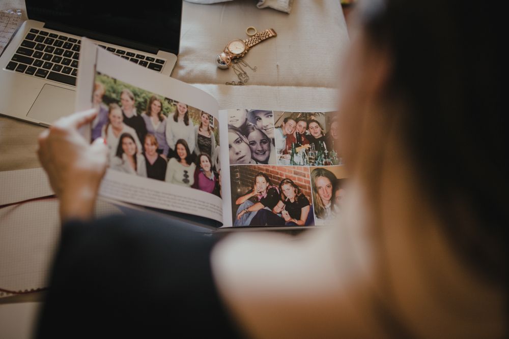 Hands hold open photo album showing group pictures on desk with laptop.