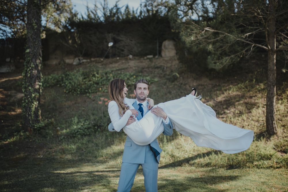 Groom in light blue suit carrying bride in white dress through wooded outdoor setting.