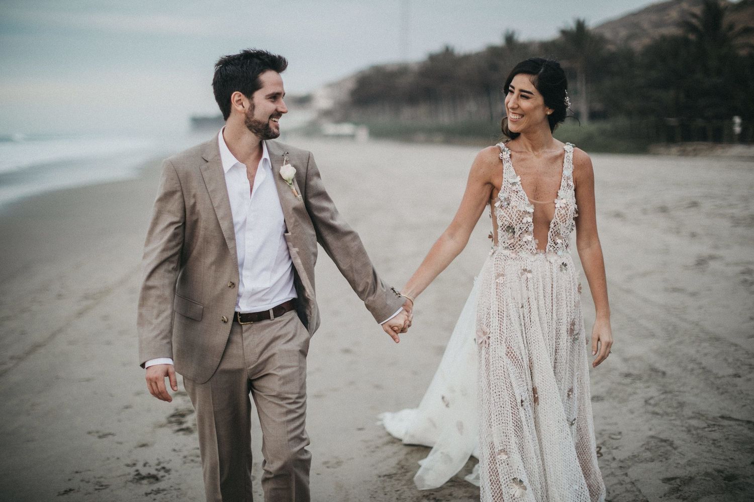 A couple walking hand in hand on a beach, with the bride in a lace gown and groom in a tan suit.