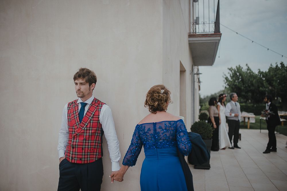 A couple in formal attire hold hands while standing against a white wall with onlookers in the background.