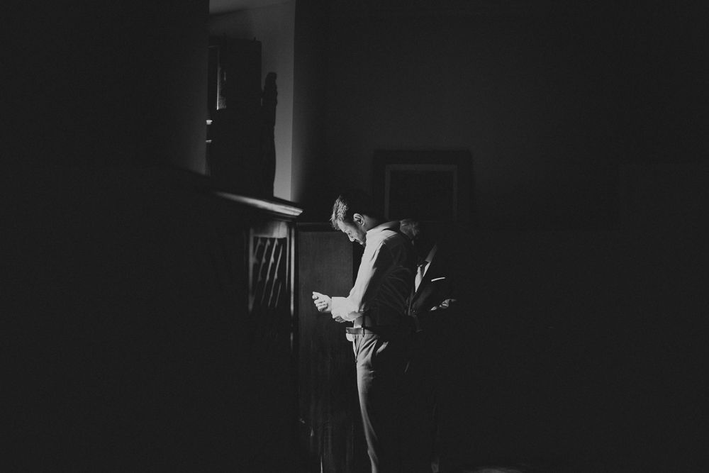 A silhouetted figure adjusts their tie in a dramatic dark room with moody lighting.