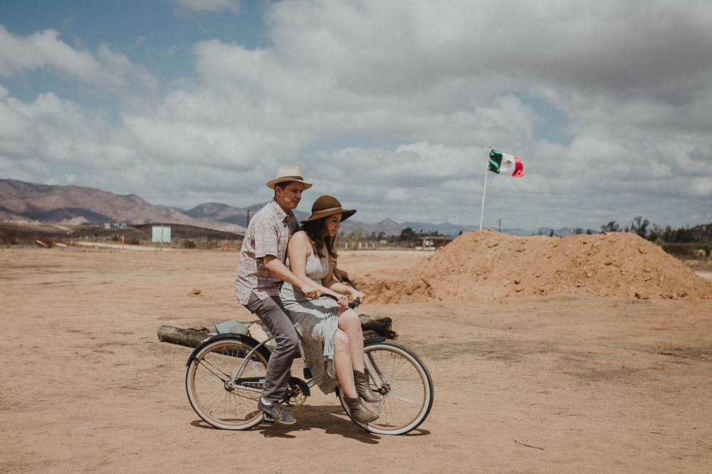 A couple rides together on a bicycle across a dusty desert landscape with a Mexican flag in the background.