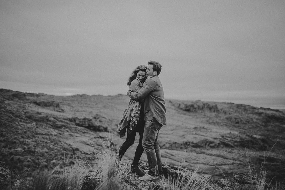 A couple embraces passionately on a windswept hilltop with mountains in the background.