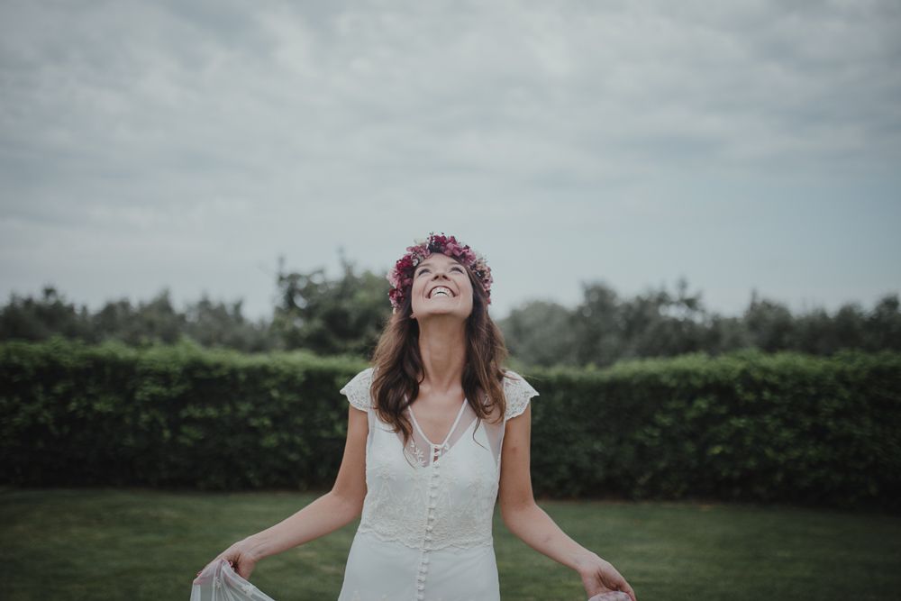 A bride in white dress and floral crown joyfully poses in garden setting.
