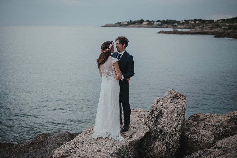 Newlyweds embrace on rocky cliff overlooking serene ocean vista.