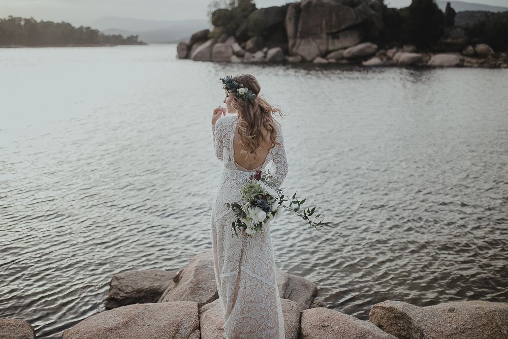 A bride in a lace wedding dress holds a bouquet while standing on rocky shoreline at dusk.
