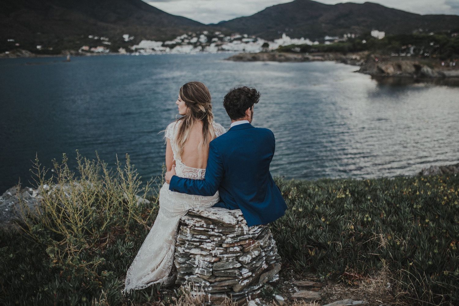 Couple sitting on stone wall overlooking Mediterranean coastal village and bay.