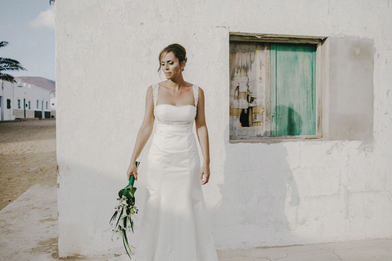 Person in white wedding dress standing beside rustic wall with turquoise window.