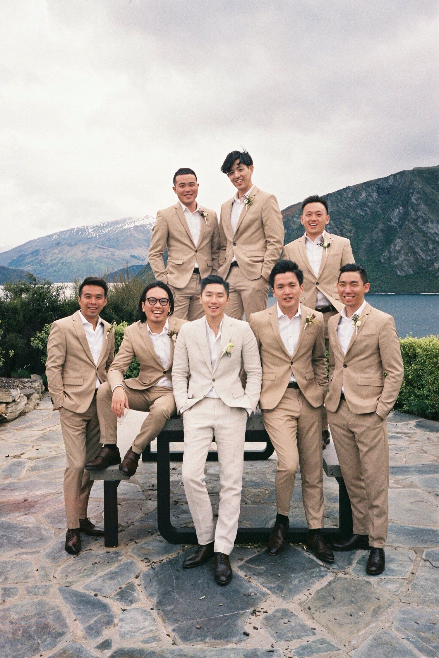 Groomsmen in coordinated beige and white suits pose together on a stone patio with mountain backdrop.