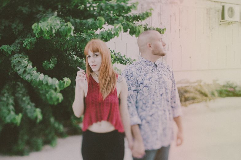 A couple stands back to back in a casual urban setting with greenery.