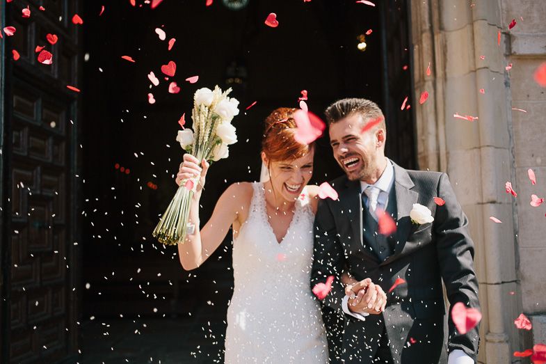 Newlyweds celebrate with red rose petals being thrown outside a stone building.