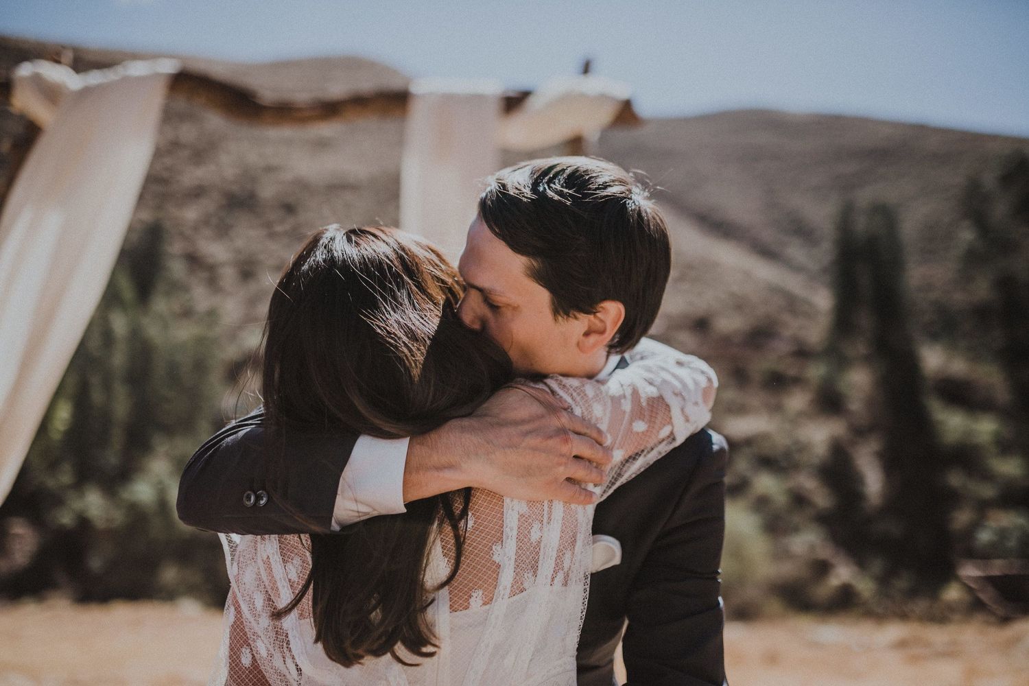 A romantic couple embraces and kisses in an outdoor mountain desert setting with flowing white fabric in the background.