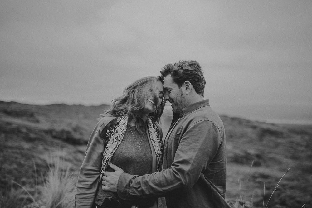 A couple shares an intimate black and white moment on a windswept hilltop.