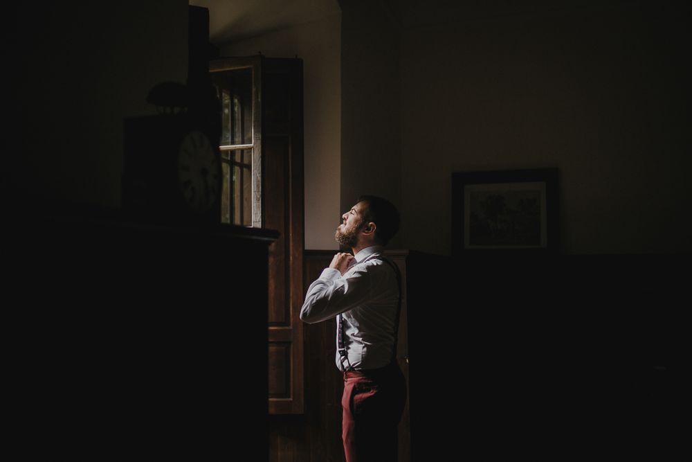 Figure in dress shirt and suspenders stands near window in shadowy room.