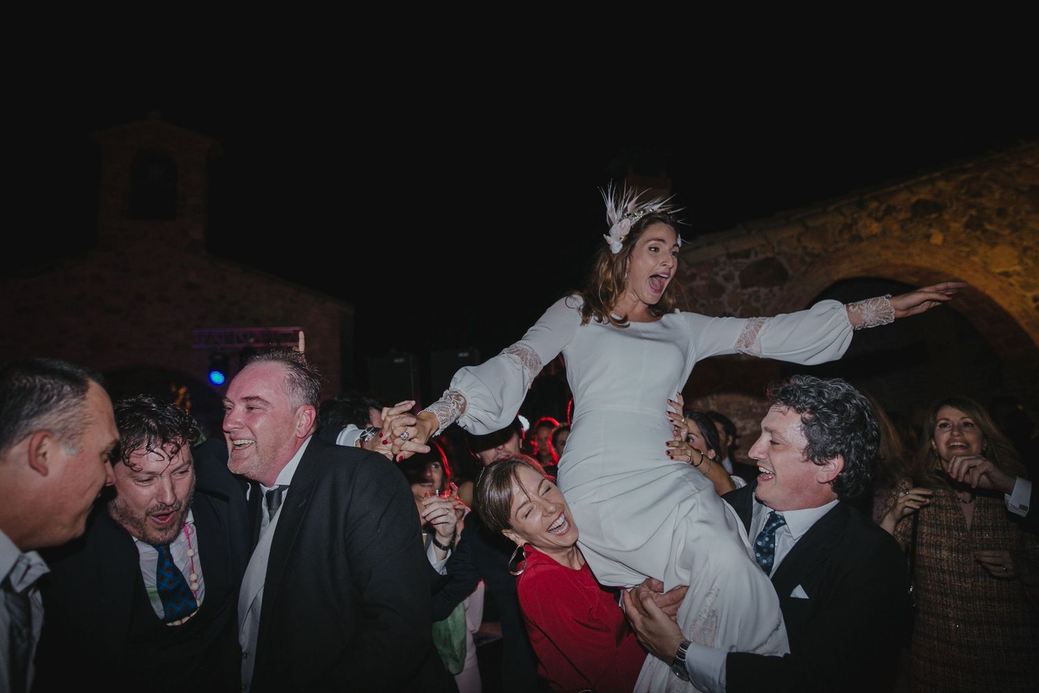 Bride being lifted by wedding guests during lively reception dance.