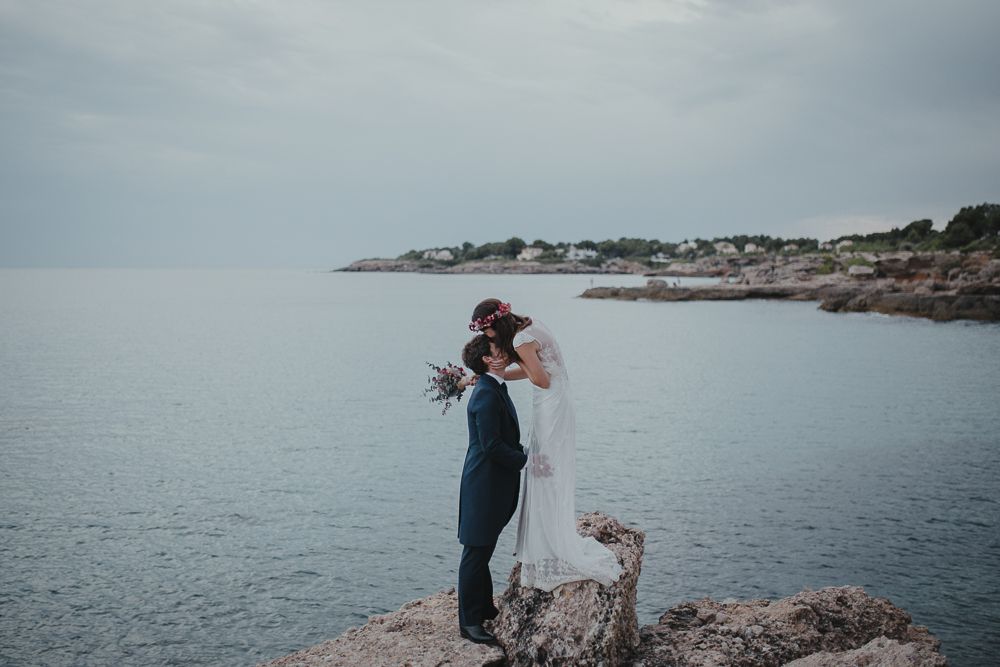Wedding couple embracing on rocky coastline with ocean town vista in background.
