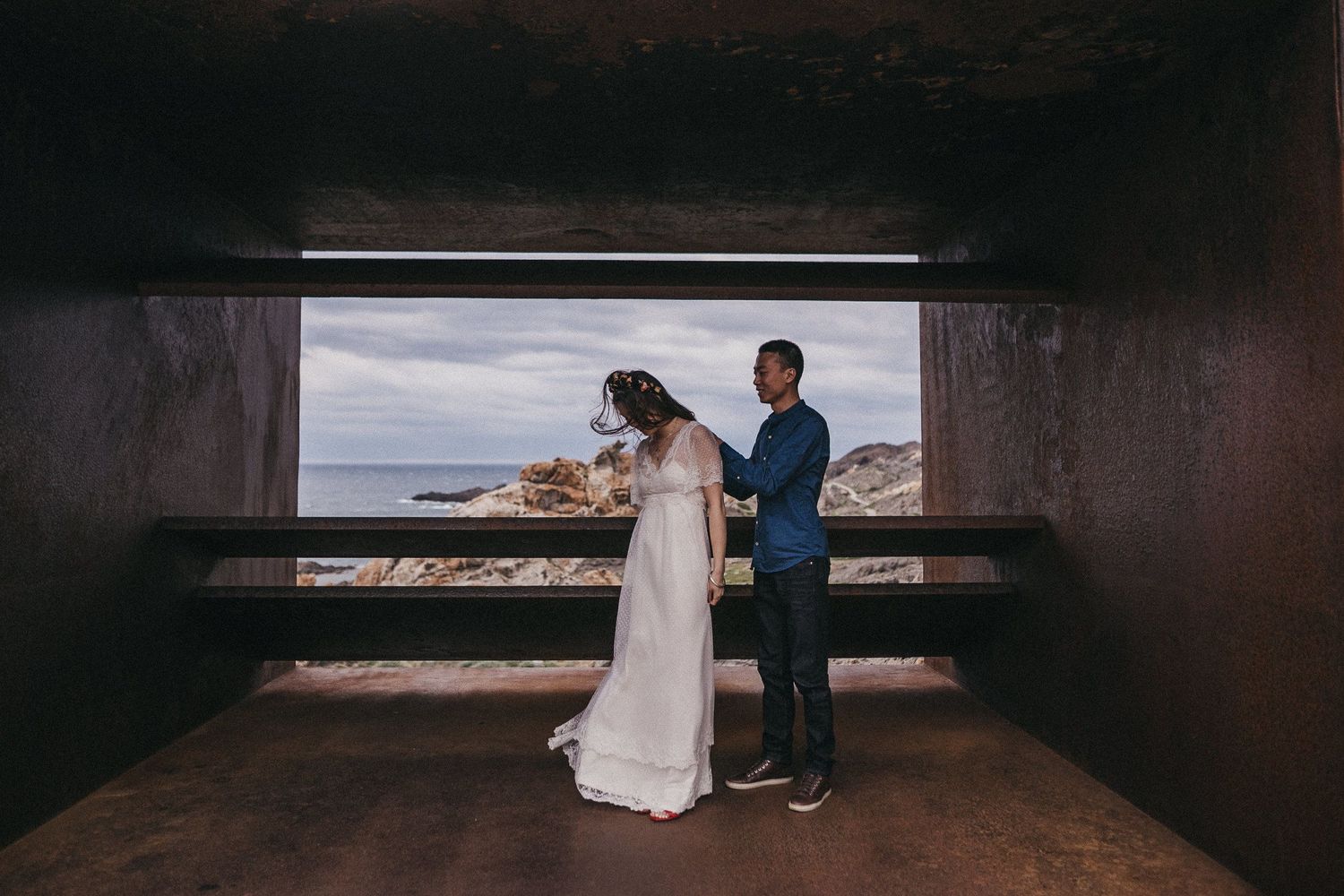 A couple in wedding attire embrace while framed by a concrete window overlooking a dramatic coastal landscape.