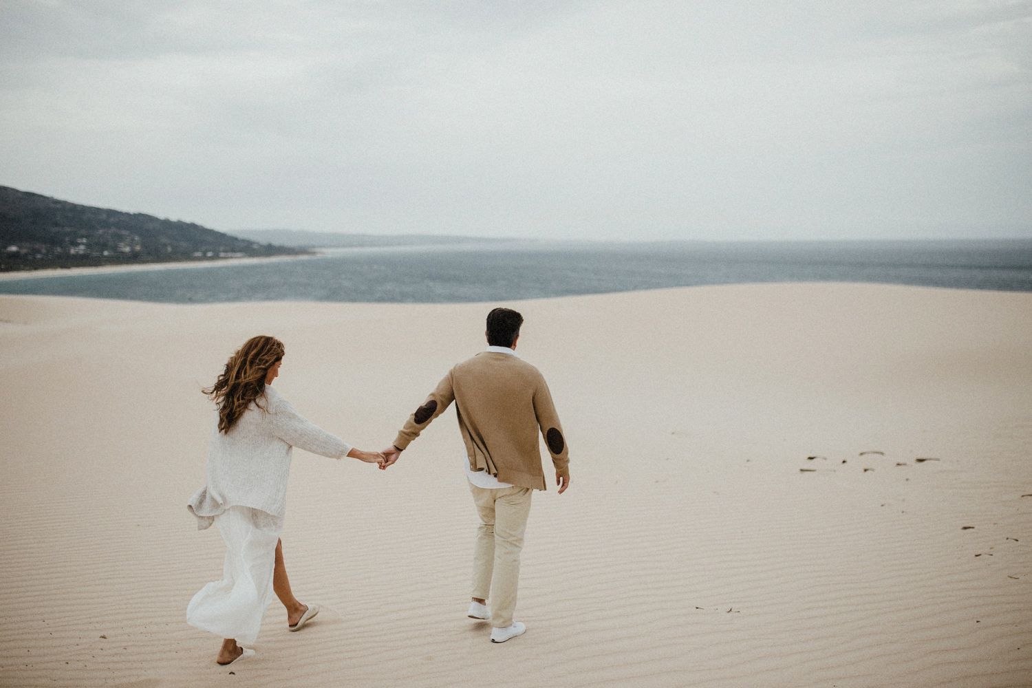 Couple walking hand in hand across vast sand dunes with ocean view in background.