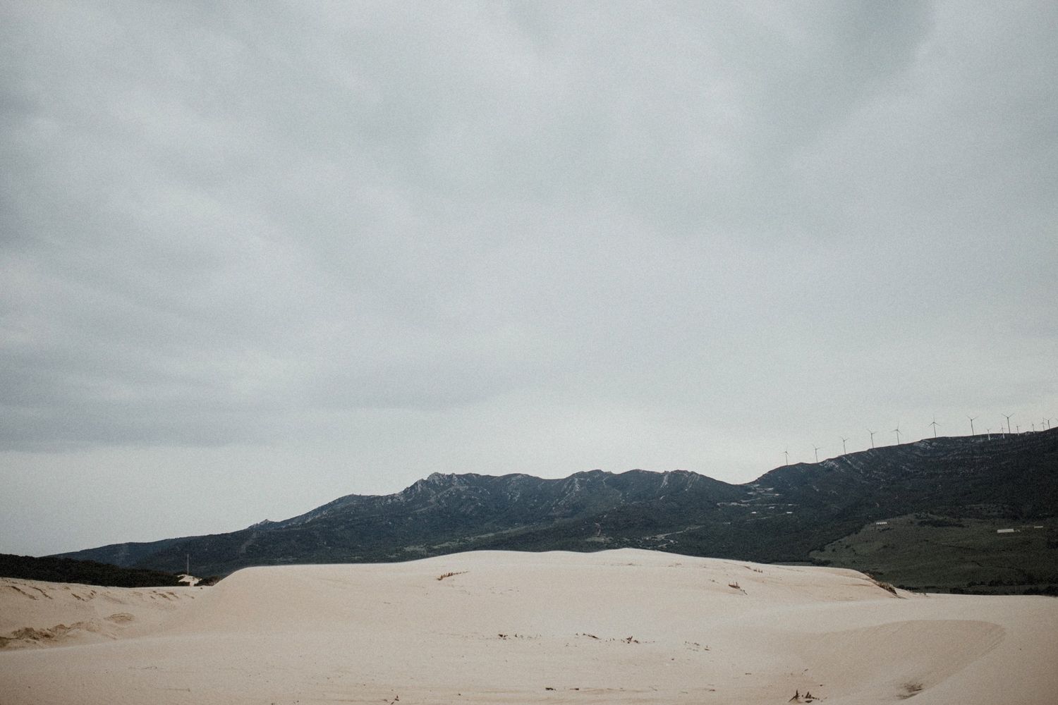 Scenic landscape of sand dunes against mountain backdrop under cloudy skies.