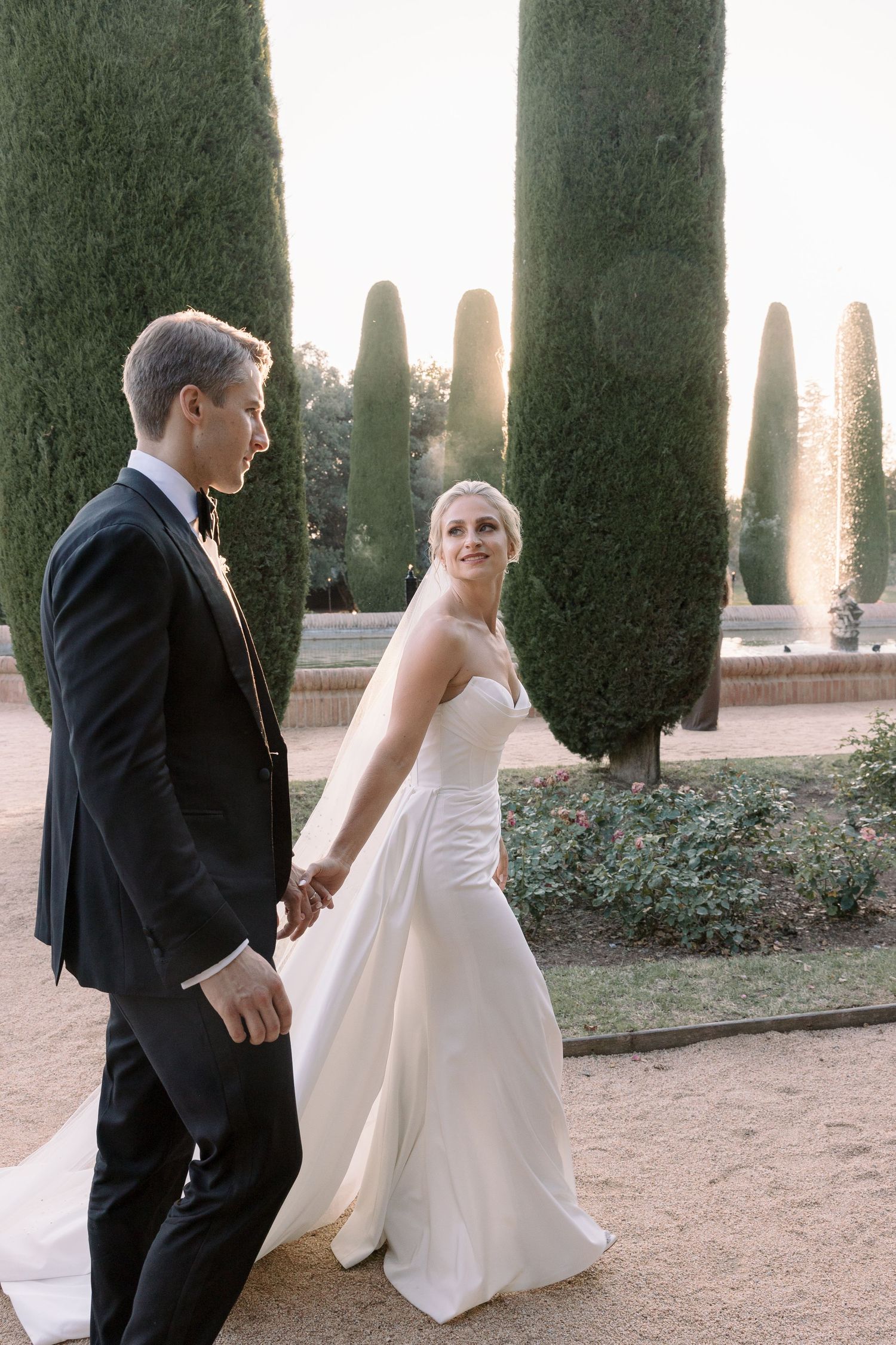 Elegant wedding couple walking through a garden with tall cypress trees at sunset.