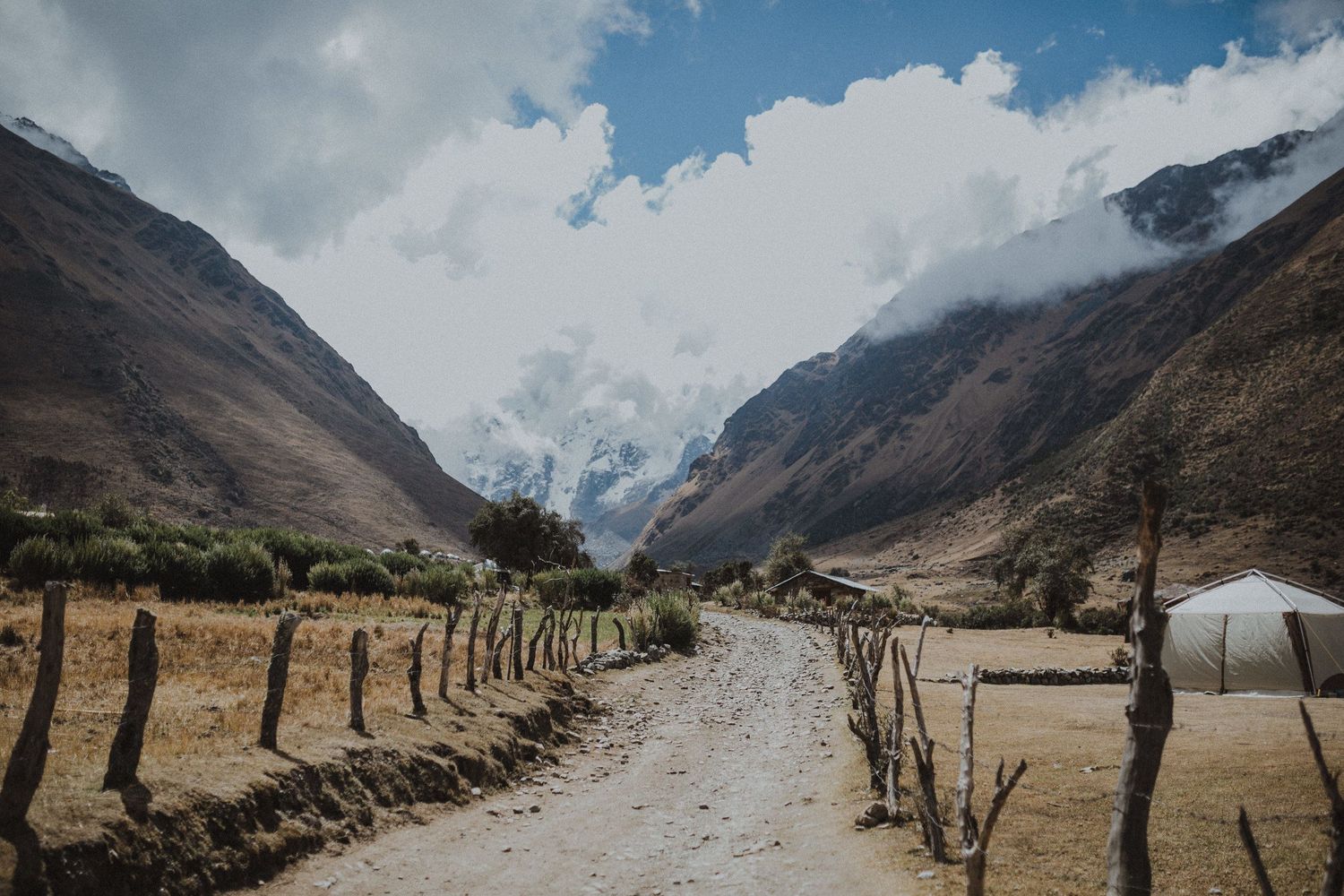 Dirt path through mountain valley with wooden fence posts and snow-capped peaks in distance.