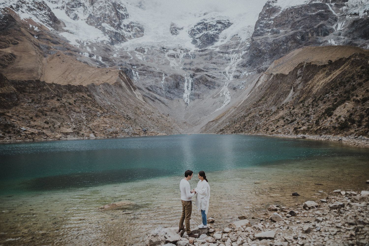 Two people standing at the edge of a turquoise mountain lake surrounded by snowy peaks.