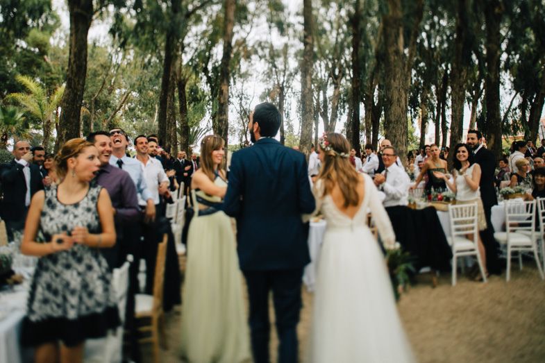 An outdoor wedding ceremony taking place under tall trees with guests seated on white chairs.